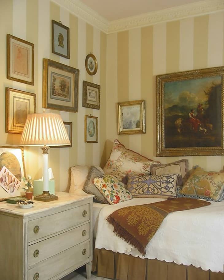 Cozy bedroom corner with striped walls, vintage art, a lamp, and a bed adorned with patterned pillows and a throw blanket.