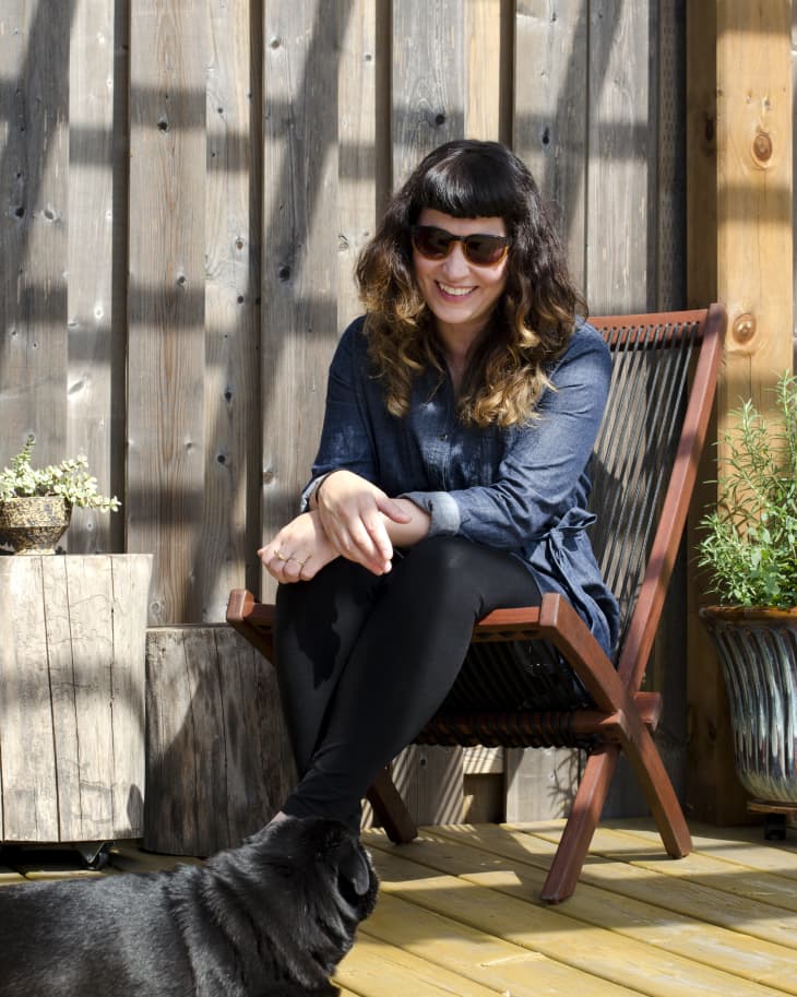 Woman in sunglasses sitting on a wooden chair on a deck, smiling at a black dog lying on the floor.