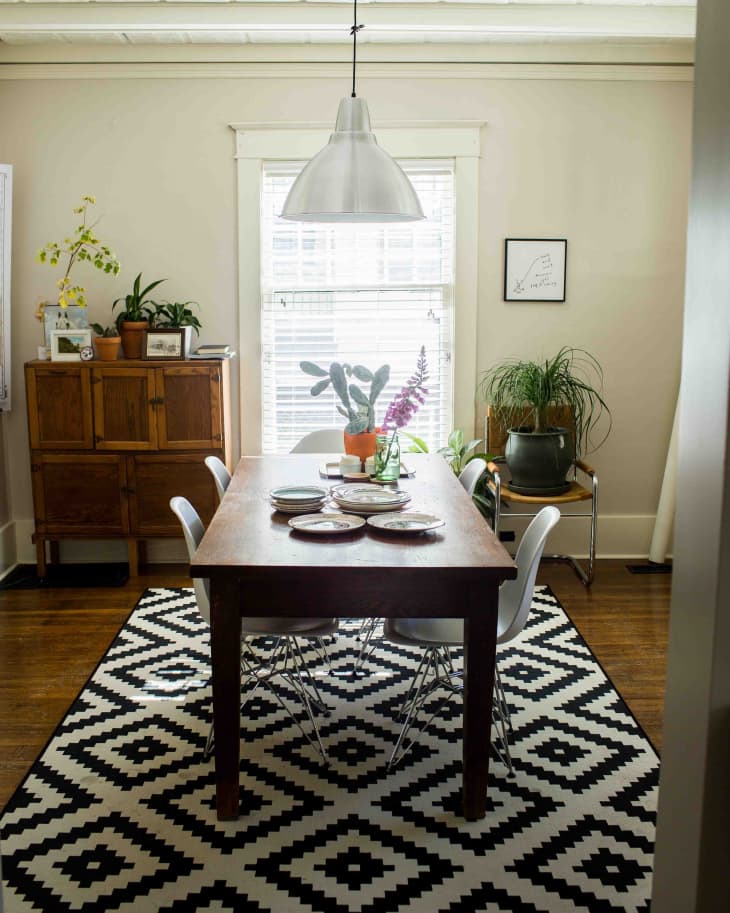 Dining room with wooden table, white chairs, geometric rug, pendant light, and plants on a sideboard.