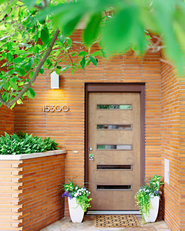 Modern wooden front door with horizontal glass panels, surrounded by brick walls, potted plants, and house number 15300.