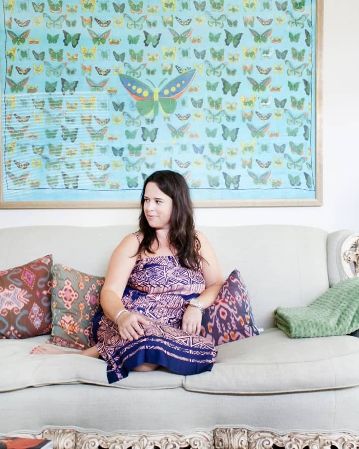 Woman in patterned dress sitting on ornate sofa with colorful pillows, large butterfly artwork on wall behind.