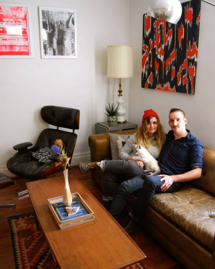 Couple sitting on a brown leather sofa with a cat, surrounded by eclectic decor, including abstract art and a vintage chair.