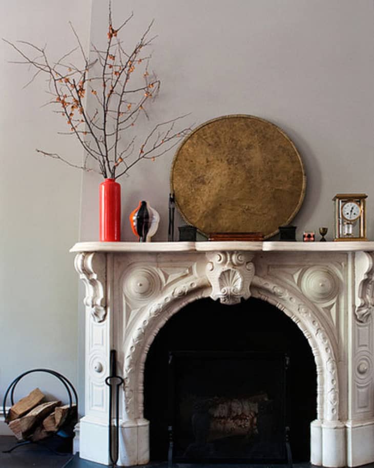 Ornate white fireplace mantel with a red vase, bare branches, a round gold tray, and a small clock.