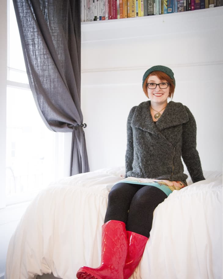 Woman in red boots sitting on a bed, wearing a gray sweater and green beanie, with books on a shelf above.