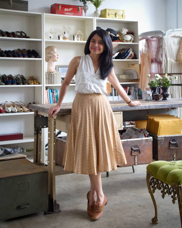 Woman in a vintage store wearing a white top and patterned skirt, standing by a wooden table with shelves of shoes and hats.