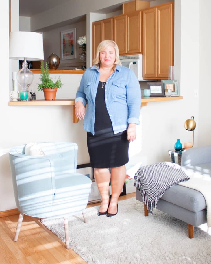 Woman in a denim shirt and black dress standing in a living room with light blue chair, gray sofa, and wooden cabinets.
