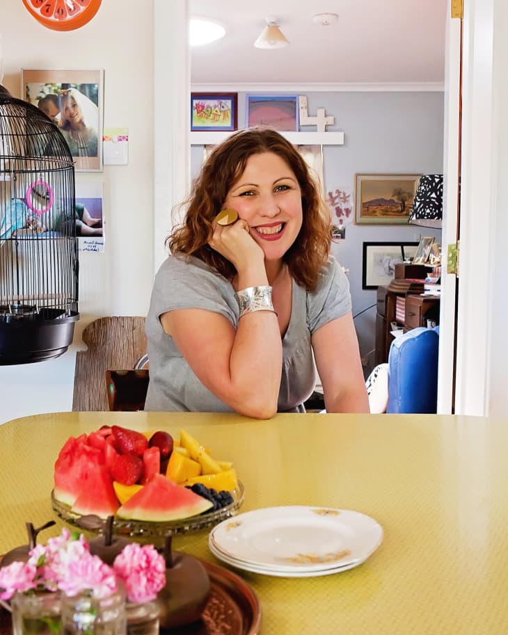 Woman smiling at a yellow table with a fruit platter, pink flowers, and a birdcage in a colorful room.