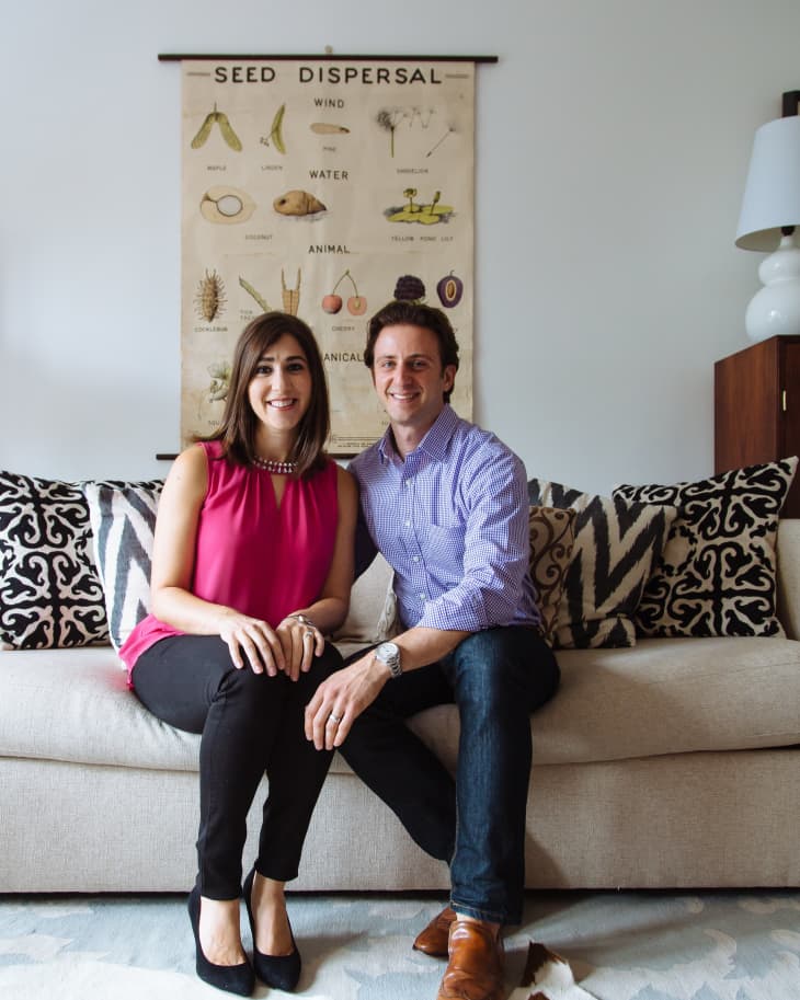 Couple sitting on a beige sofa with patterned cushions, under a seed dispersal poster, next to a white lamp.