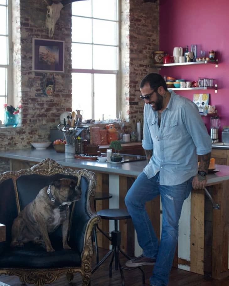 Man in denim outfit leaning on kitchen counter, looking at a dog sitting on an ornate chair in a rustic kitchen.