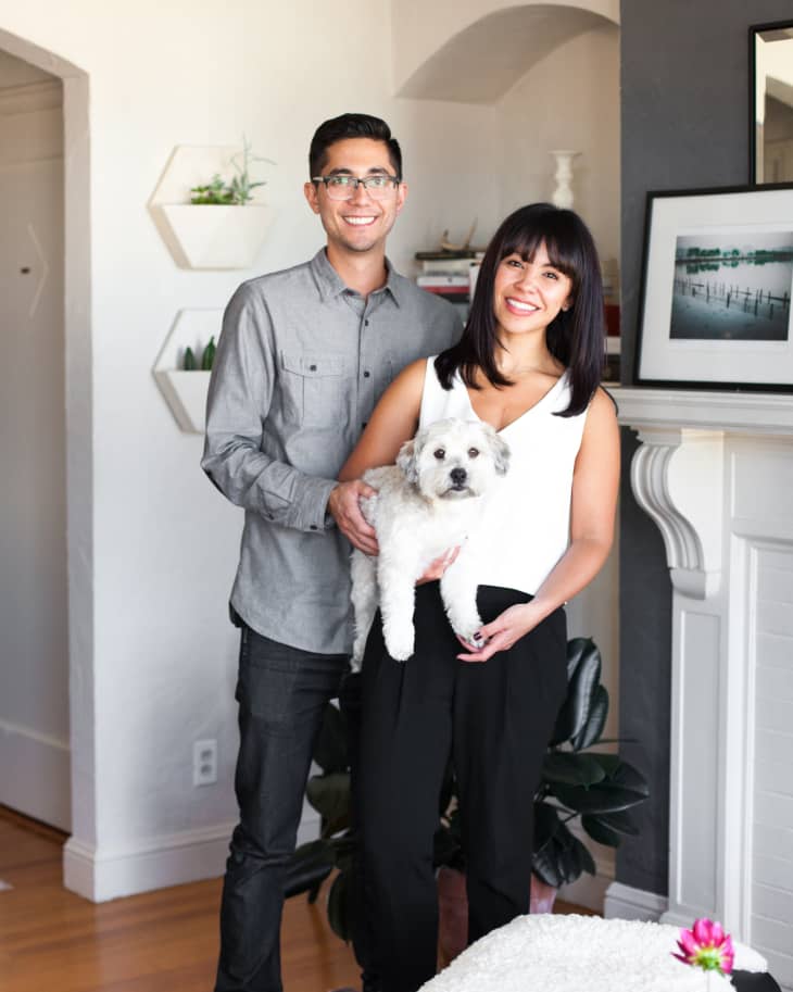 Couple smiling in living room holding a small white dog, with wall art and plants in the background.