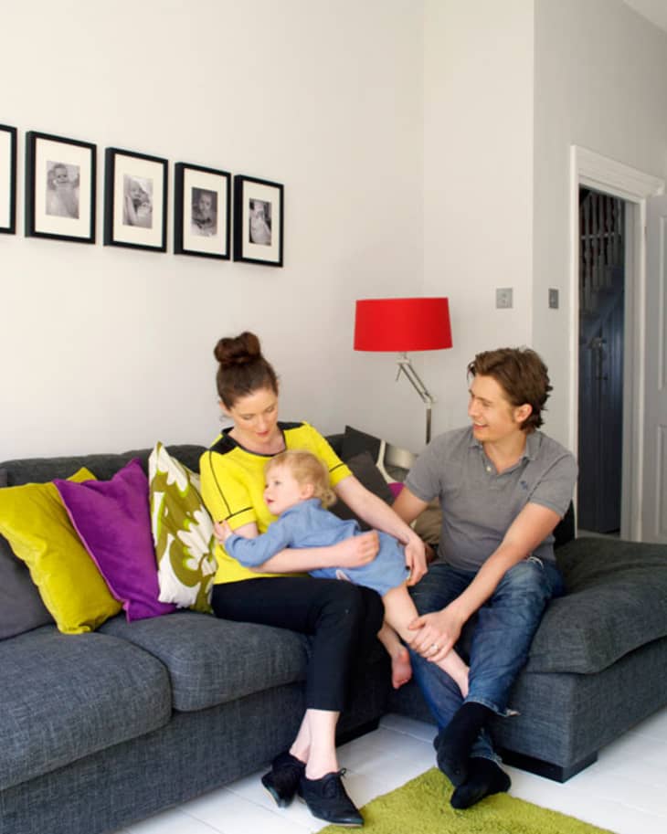 Family sitting on a gray sofa with colorful cushions, red lamp, and framed photos on the wall.