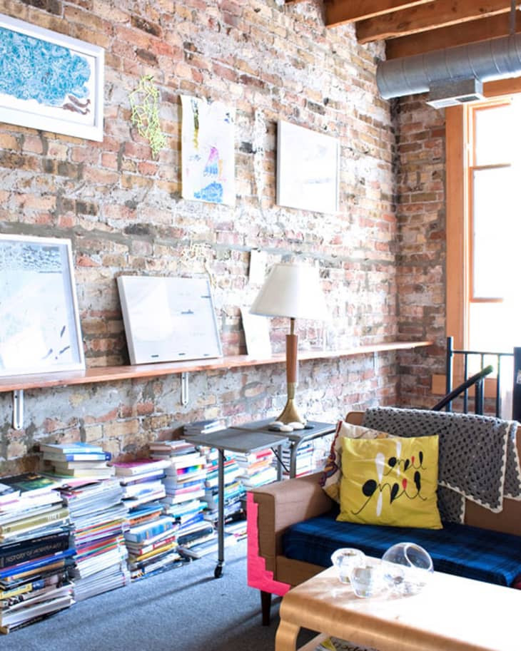Cozy living room with exposed brick wall, art, books, a blue sofa, and a yellow pillow.