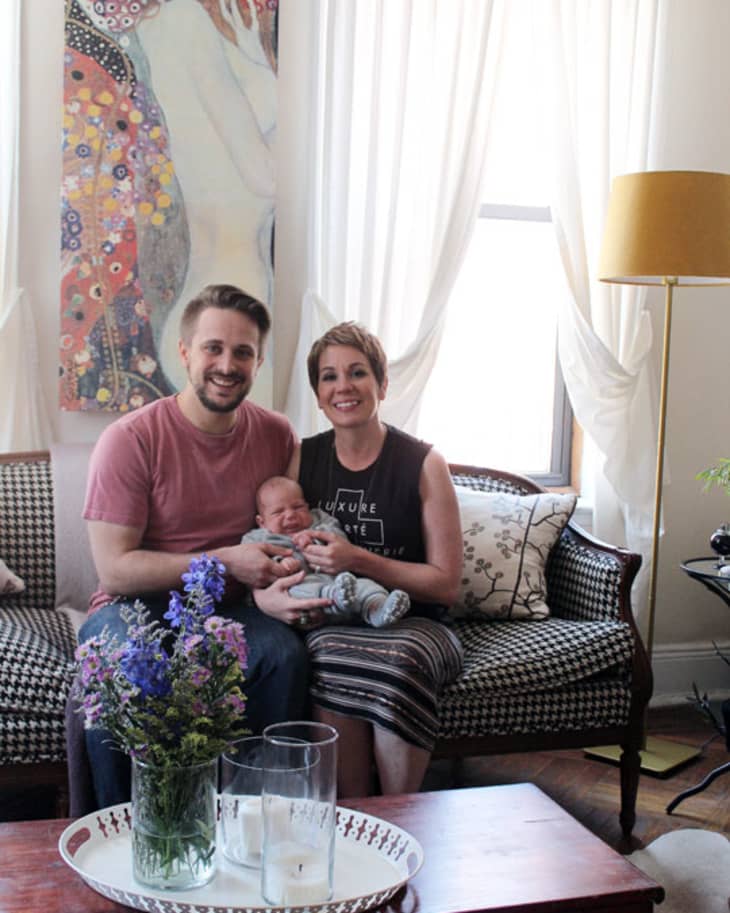 Couple sitting on a patterned sofa with a baby, under a Klimt-style painting, next to a wooden table with flowers.