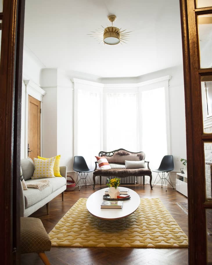 Living room with a vintage sofa, modern chairs, yellow rug, and oval coffee table with books and flowers.