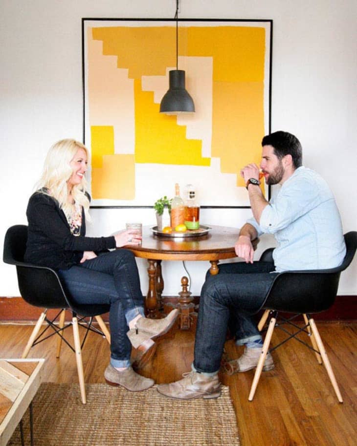 Two people sitting at a round table with drinks, under a black pendant light, in front of a yellow geometric painting.