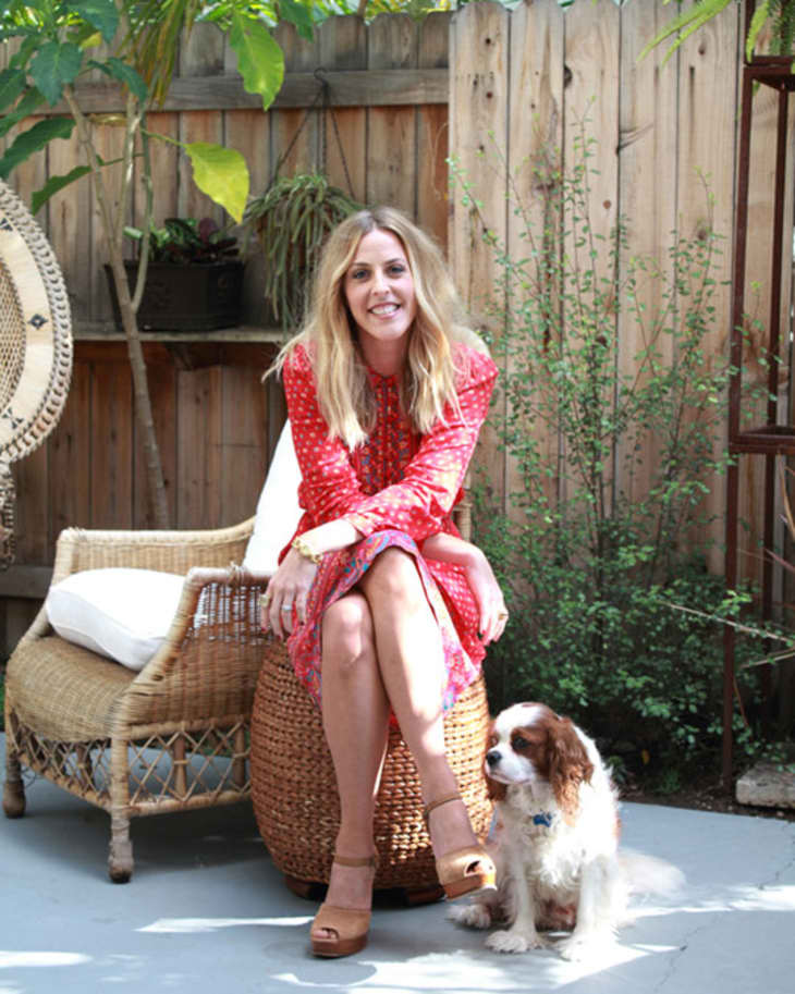 Woman in a red dress sitting on a wicker chair in a garden with a small dog beside her.