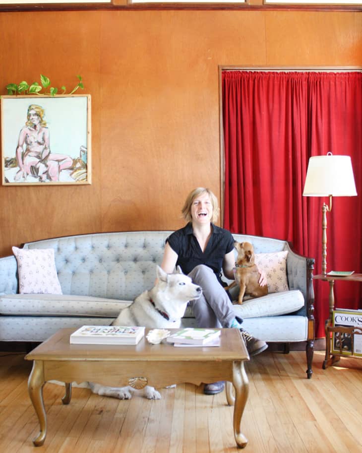 Woman sitting on a blue sofa with two dogs, wooden coffee table, red curtains, and a painting on the wall.