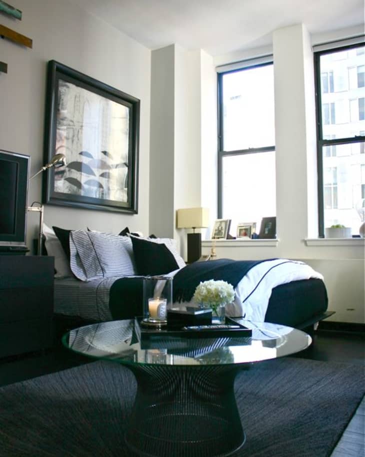 Modern bedroom with a glass coffee table, black and white bedding, large artwork, and framed photos on the windowsill.