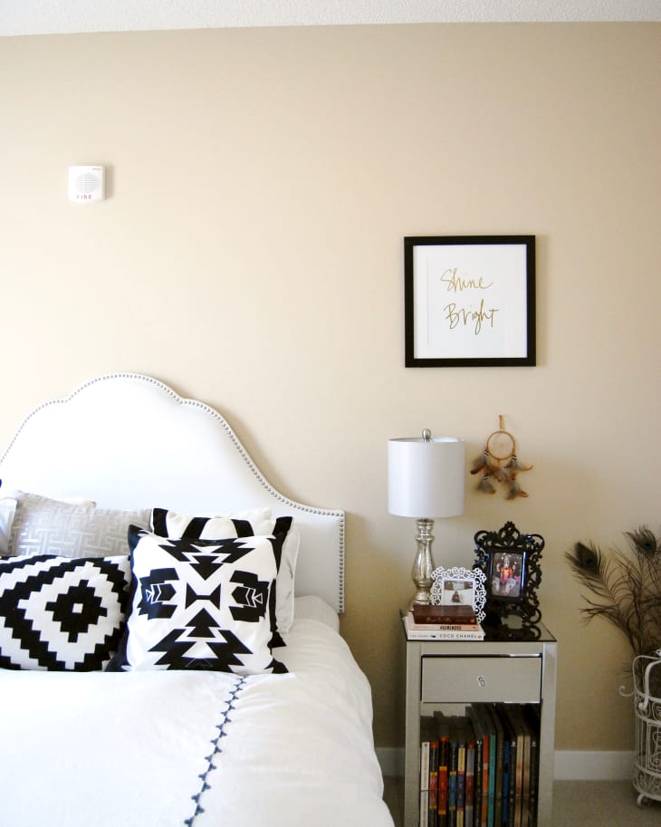 Bedroom with white bed, black and white geometric pillows, mirrored nightstand, lamp, framed art, and dreamcatcher.