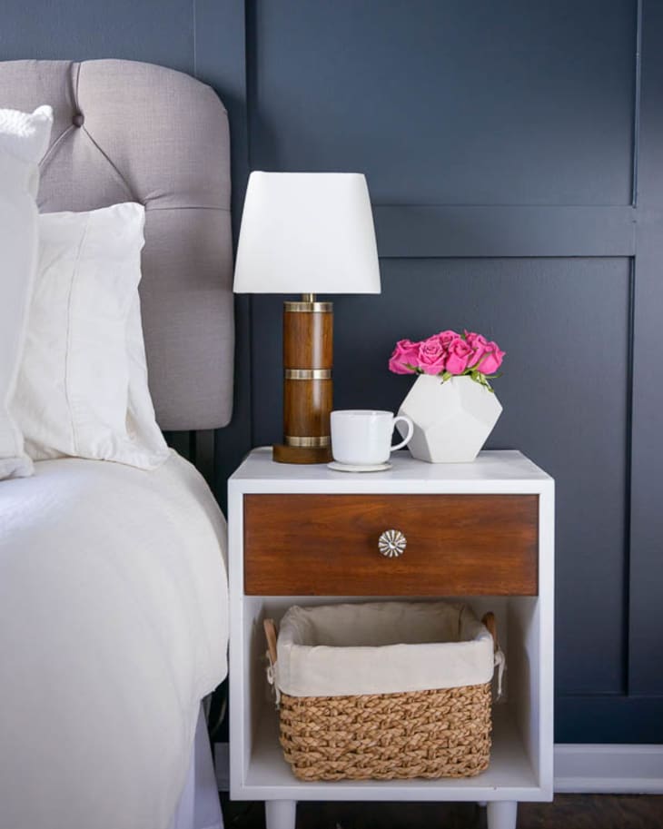 Bedside table with lamp, white mug, pink roses in vase, and wicker basket below against dark wall.
