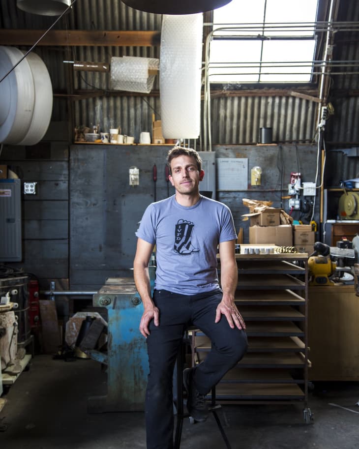 Man in a workshop sitting on a stool, wearing a graphic t-shirt, surrounded by tools and industrial equipment.