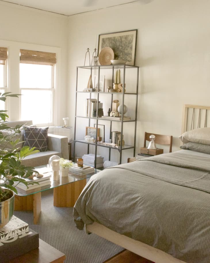 Cozy bedroom with a gray bedspread, glass coffee table, potted plant, and a metal bookshelf filled with decor and books.