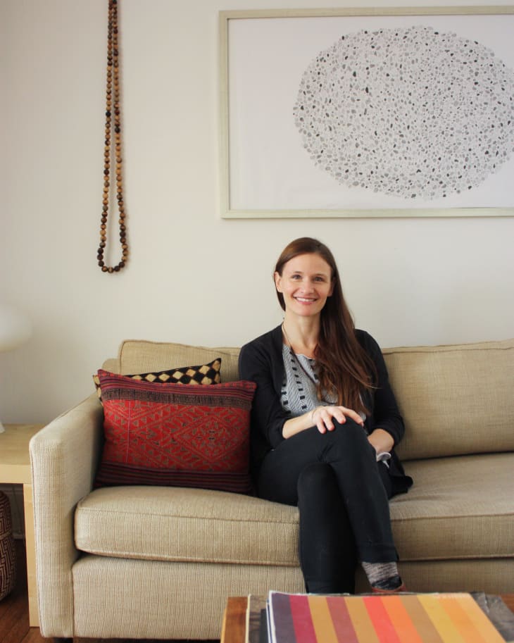 Woman sitting on beige sofa with red patterned cushions, abstract wall art, and a wooden coffee table with books.