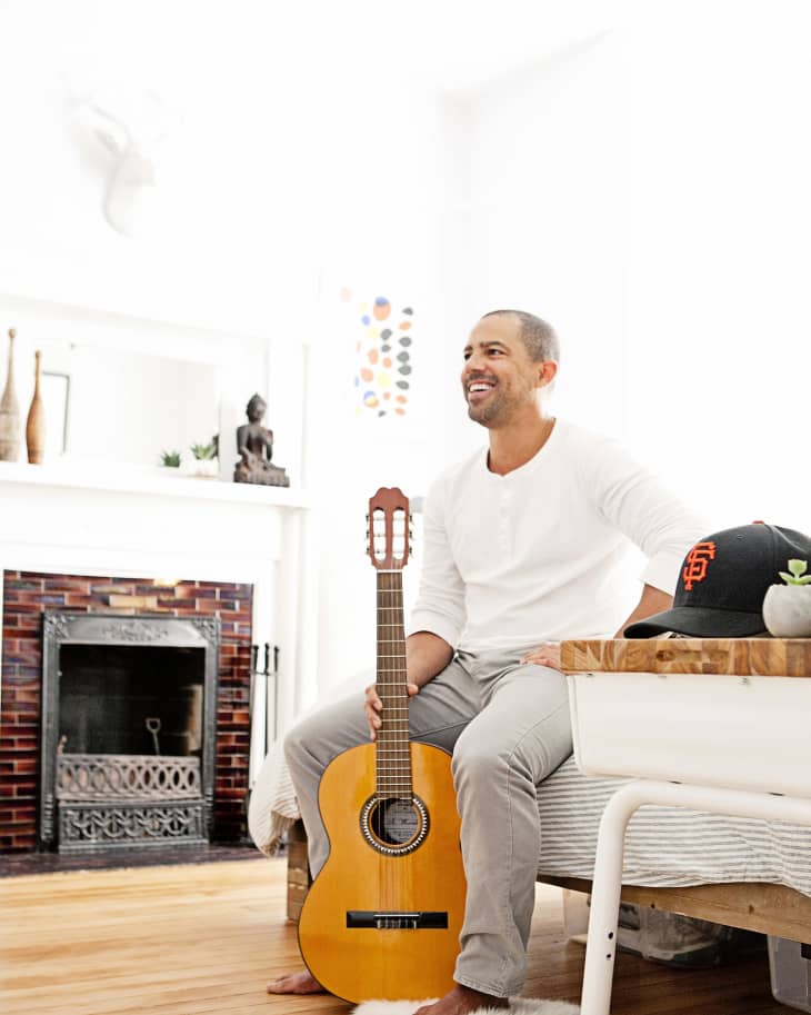 Man sitting on bed holding a guitar, near a fireplace with decor and a cap on the table.