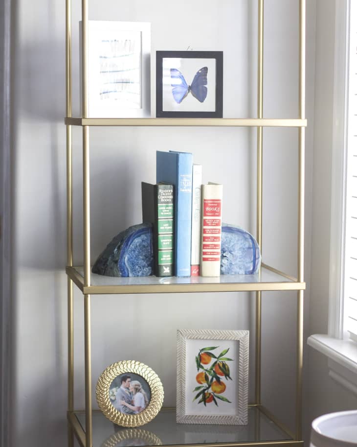 Gold shelving unit with books, framed butterfly, abstract art, and decorative geode. Lower shelf has framed photo and orange print.
