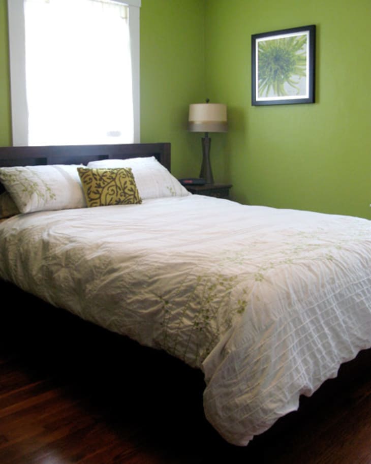 Bedroom with green walls, white bedding, decorative pillow, framed plant artwork, and a bedside lamp.