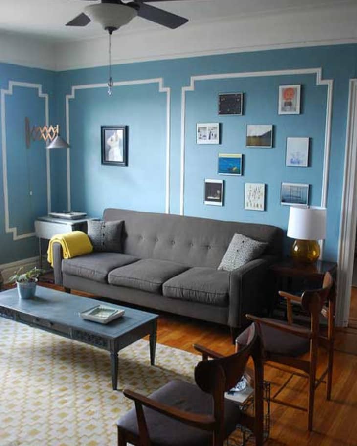 Living room with blue walls, gray sofa, gallery wall art, wooden chairs, and a yellow throw blanket.