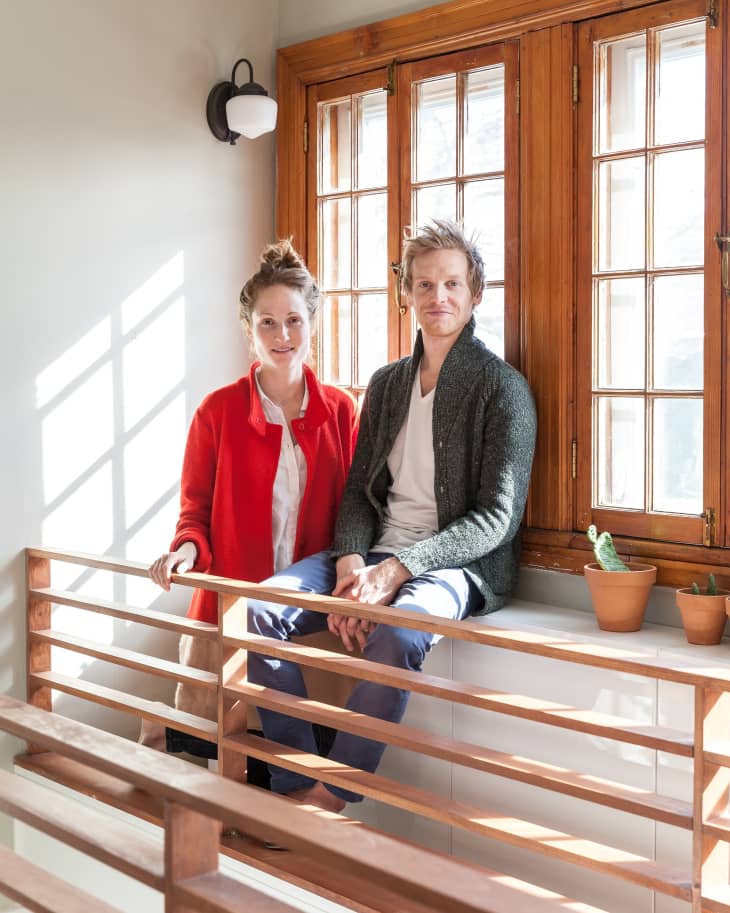Two people sitting on a wooden window ledge with potted plants, sunlight streaming through the window.