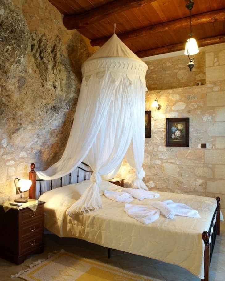 Rustic bedroom with stone walls, wooden ceiling, canopy bed, yellow bedding, and a bedside table with a lamp.