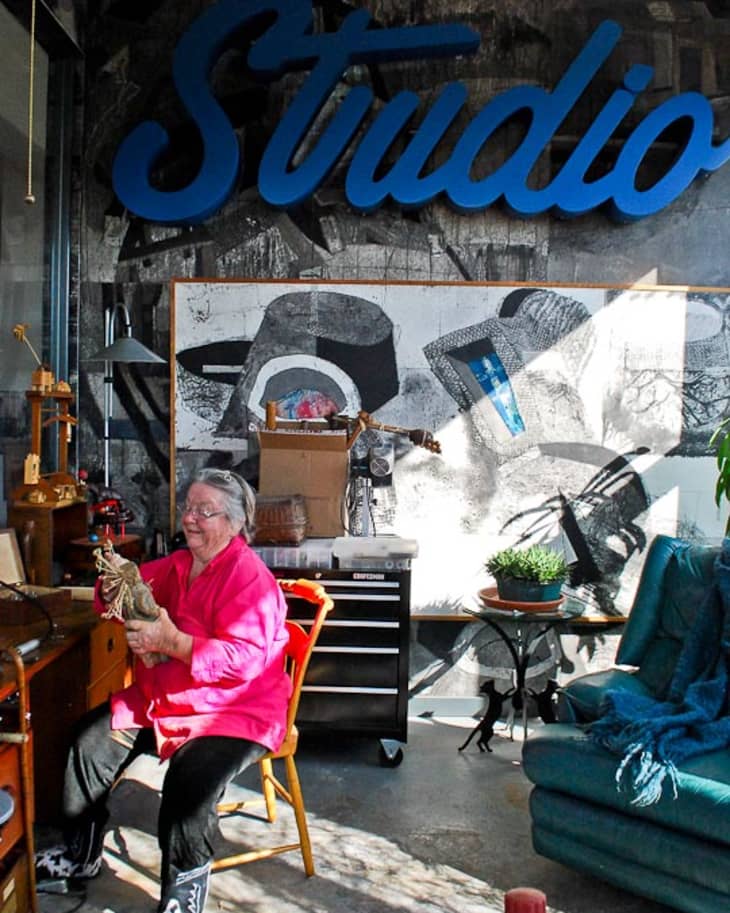 Woman in a pink shirt crafting in an art studio with a large "Studio" sign and abstract wall art.