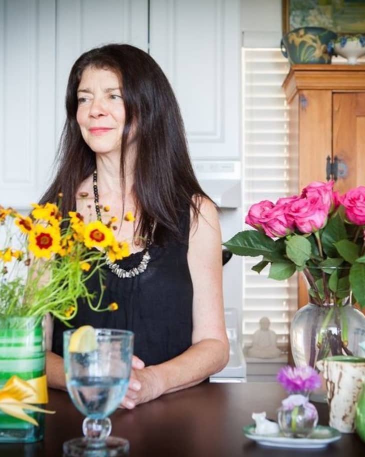 Woman in black dress at table with yellow and pink flowers, glass of water, and wooden cabinet in background.