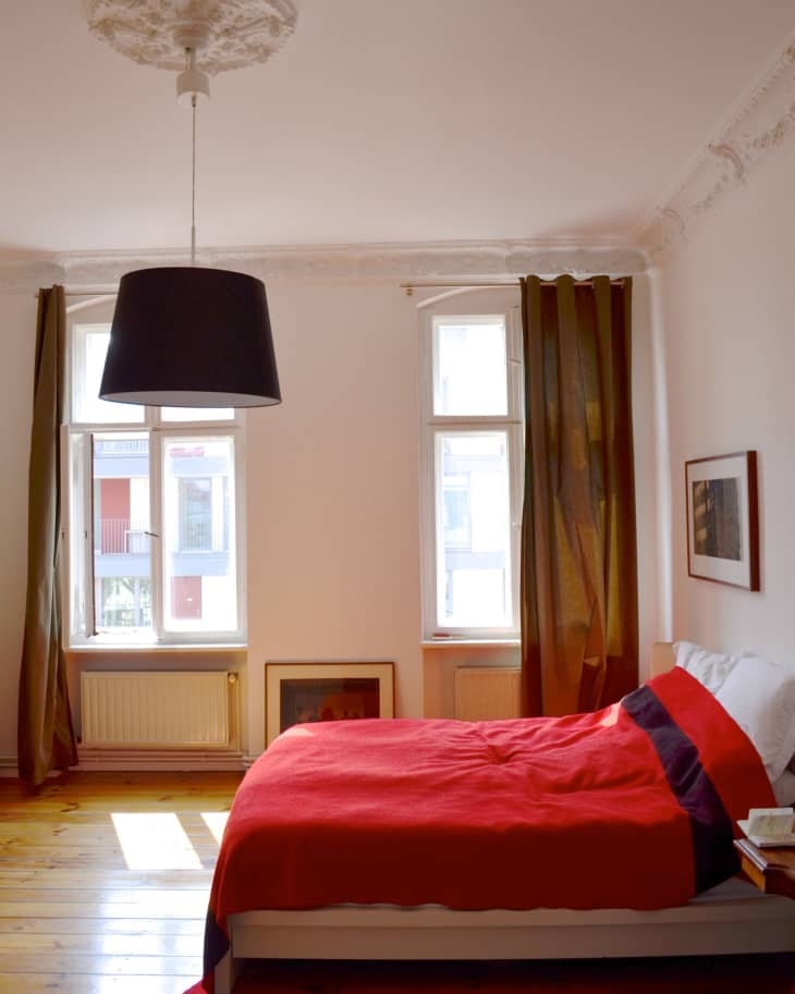 Bedroom with red bedding, wooden floor, large windows, green curtains, and a black pendant light.