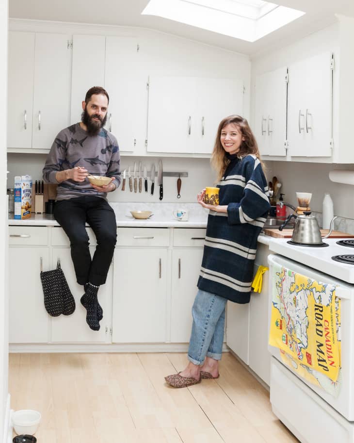 Two people in a white kitchen, one sitting on the counter, both holding bowls, with a map towel on the oven.