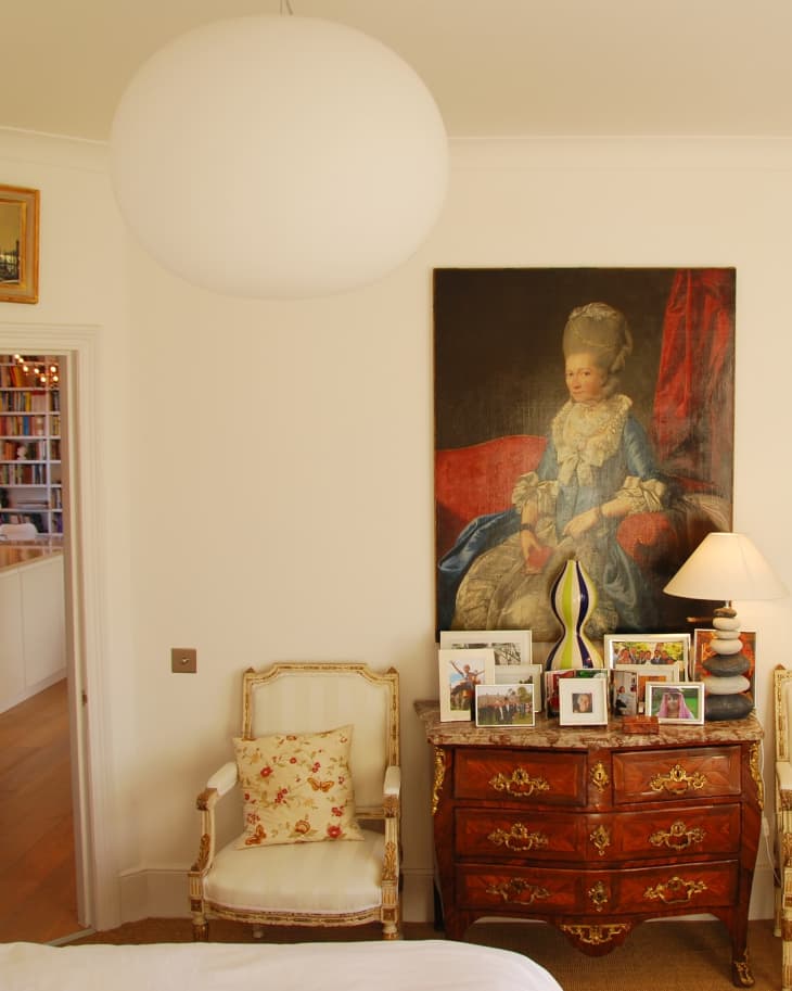 Elegant room with ornate chair, floral cushion, vintage dresser, framed photos, and a large portrait on the wall.