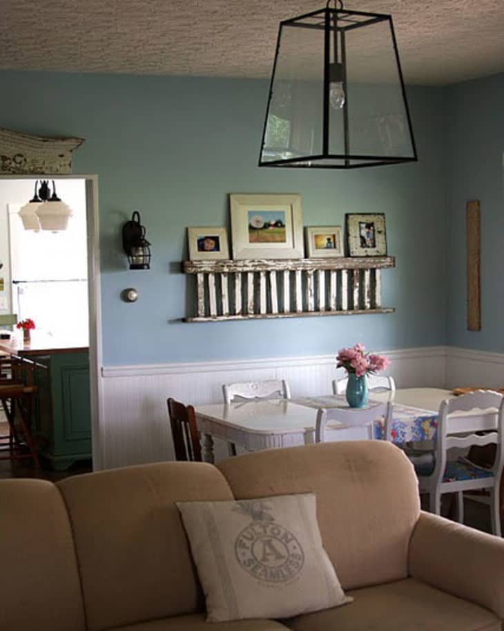 Living room with beige sofa, blue walls, white dining table, chairs, and framed art on shelf. Large pendant light above.
