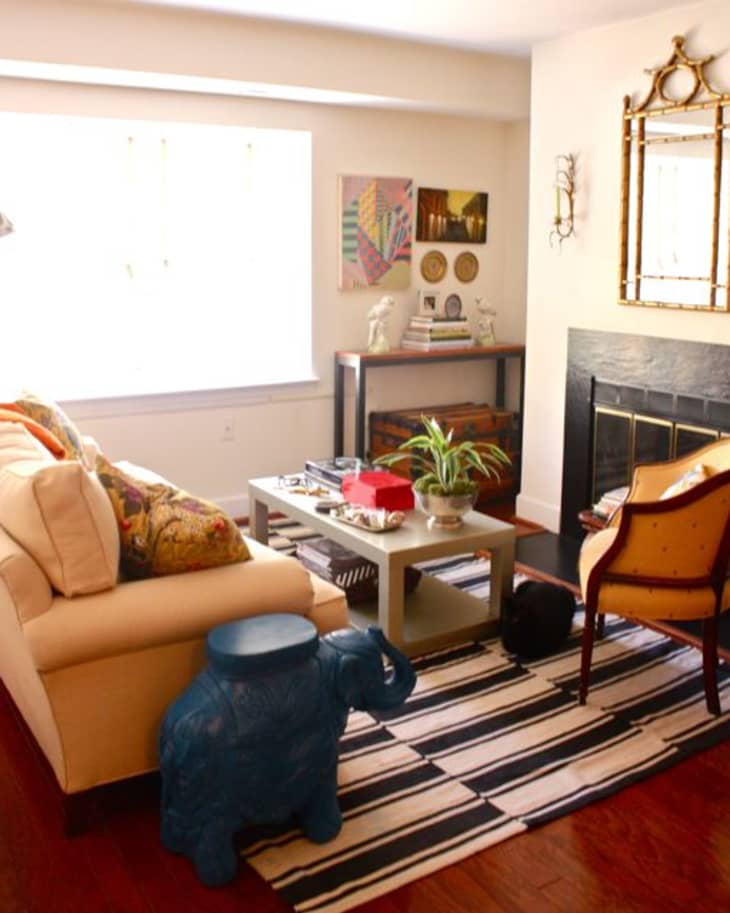 Cozy living room with a beige sofa, blue elephant stool, striped rug, and a fireplace. Decor includes plants and colorful art.
