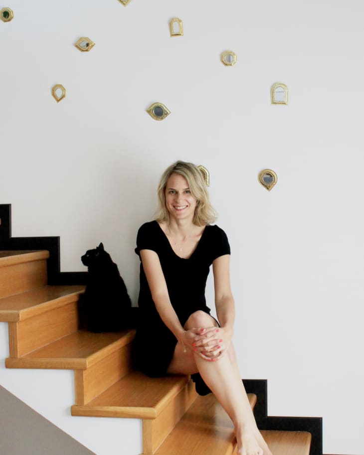 Woman in black dress sitting on wooden stairs with a black cat, surrounded by small decorative mirrors on the wall.