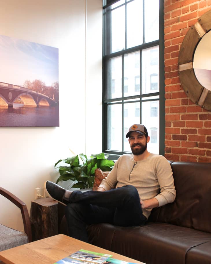 Man sitting on a brown leather sofa in a brick-walled room with a large window, plants, and a wooden coffee table with magazines.