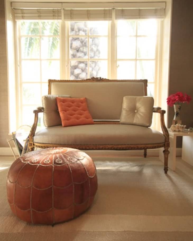 Elegant living room with a vintage sofa, orange and beige cushions, leather pouf, and a side table with red flowers.