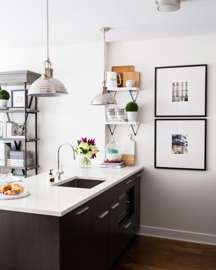 Modern kitchen with dark cabinets, white countertop, pendant lights, framed art, and shelves with decor and plants.
