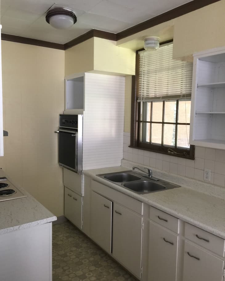 Small kitchen with beige cabinets, double sink, built-in oven, and large window with blinds.