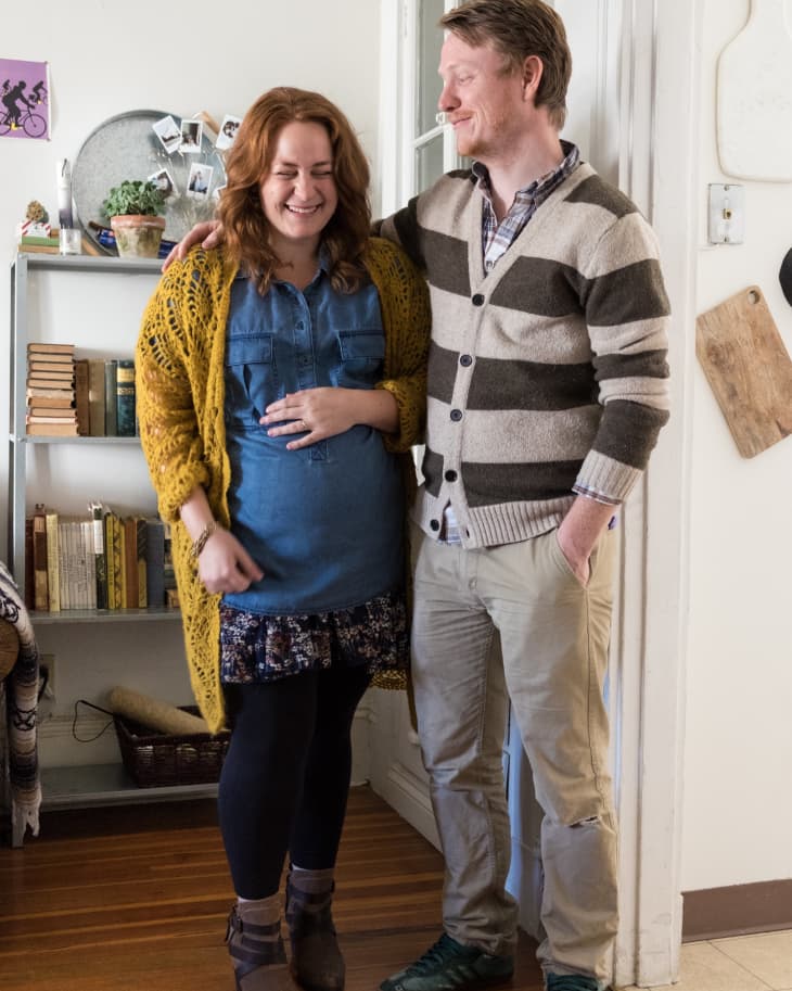 Couple smiling in a cozy room with bookshelves, wearing casual clothes; woman in a yellow cardigan, man in a striped sweater.