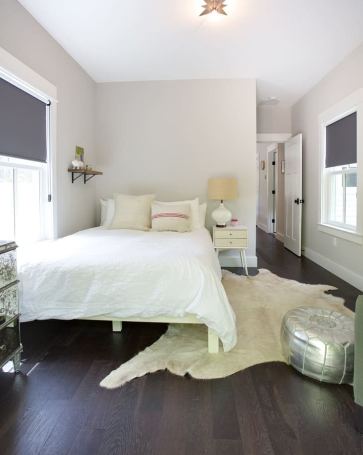 Minimalist bedroom with white bedding, dark wood floors, a fur rug, silver pouf, and a small nightstand with a lamp.