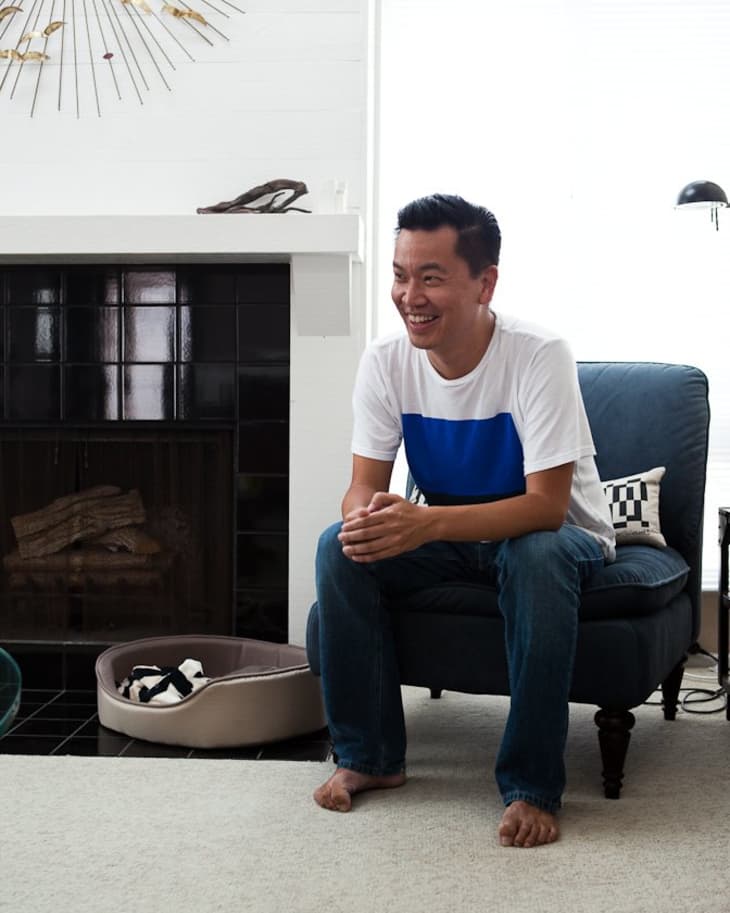 Man sitting on a blue chair in a living room with a fireplace, sunburst wall art, and a dog bed on the floor.