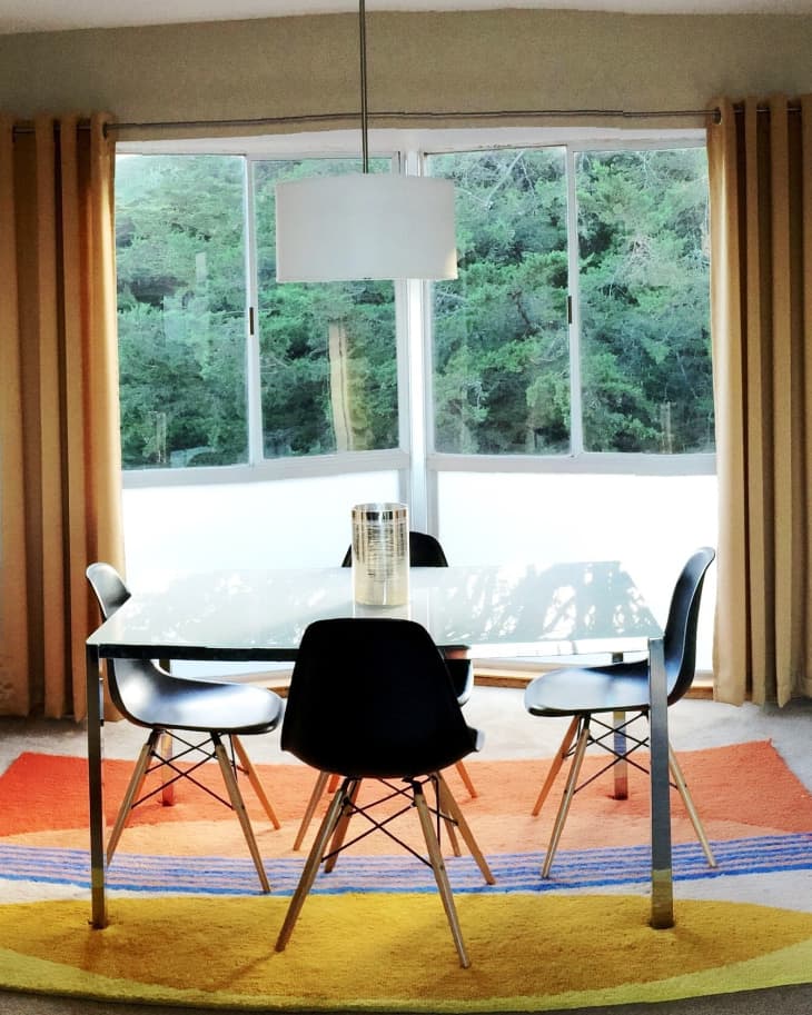 Dining area with a glass table, four black chairs, colorful rug, and large windows with forest view.