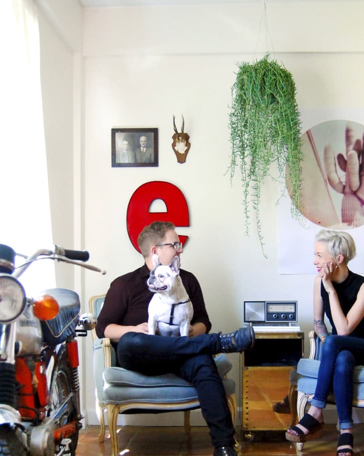 Man and woman sitting in a living room with a dog, vintage decor, red letter "e," and a hanging plant.